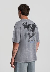 Uomo che indossa una t-shirt grigia sbiadita e oversize con una grafica di angelo nera e la scritta "Anger Faith Los Angeles" sulla parte posteriore, abbinata a jeans azzurri chiari.