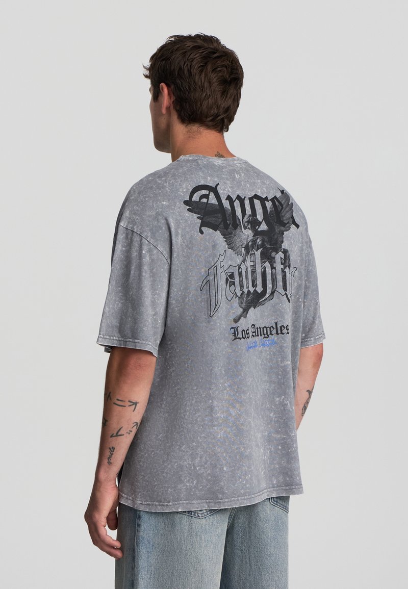 Uomo che indossa una t-shirt grigia sbiadita e oversize con una grafica di angelo nera e la scritta "Anger Faith Los Angeles" sulla parte posteriore, abbinata a jeans azzurri chiari.