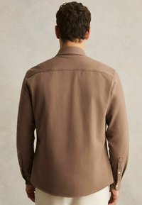 Cămașă lungă maro cu mâneci lungi, fabricată dintr-un material neted, având un guler clasic, manșete cu nasturi și un tiv rotunjit. Fără modele sau accente vizibile.
