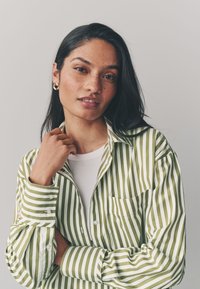 Camicia a righe verdi e bianche con bottoni e taschino sul petto, caratterizzata da una texture morbida e una vestibilità ampia, indossata sopra una maglia bianca.