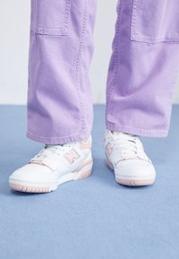Baskets blanches avec accents roses et semelles texturées, associées à un pantalon large couleur lavande, sur un fond bleu.