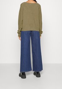 Olivgrüner, gestrickter Pullover mit gesenkten Schultern, kombiniert mit hochgeschnittenen, blauen, weit geschnittenen Jeans und schwarzen Plateauschuhen.