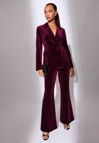 Burgonya színű bársony, dupla mellényes blézer, hozzáillő, bővülő nadrággal, zsebekkel és megkötős övvel. A modellen egy kicsi fekete kézitáska van.