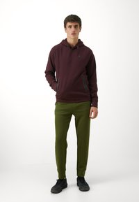 Un hoodie bordeaux avec un logo sur le devant, doté de poches zippées ; associé à un pantalon de jogging vert et à des baskets noires. Texture lisse, coupe décontractée.