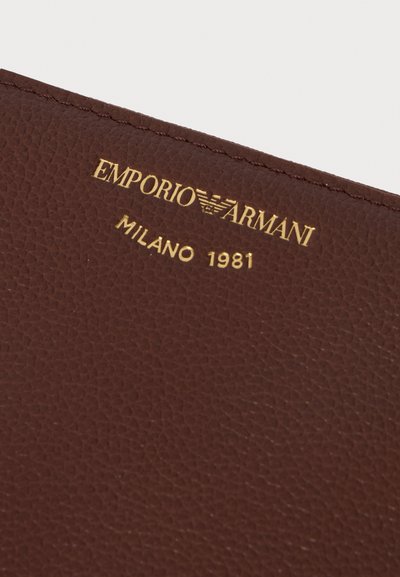 Κοντινή λήψη καφέ δέρματος με ανάγλυφο χρυσό λογότυπο Emporio Armani Milano 1981 και ραφές κατά μήκος της άκρης.
