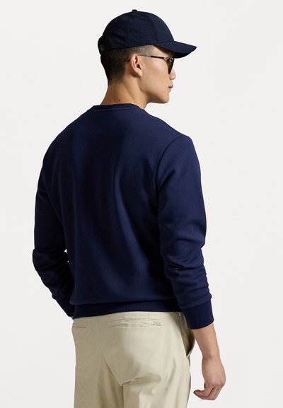 Sweatshirt bleu marine en tissu doux, avec un col rond et des poignets ribbés, porté avec un pantalon beige. Design simple et épuré.