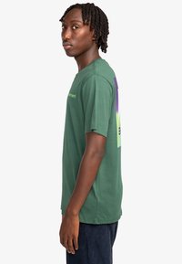 Camiseta de algodón verde con mangas cortas, que presenta un diseño gráfico en púrpura y verde en la parte posterior, y un dobladillo recto.