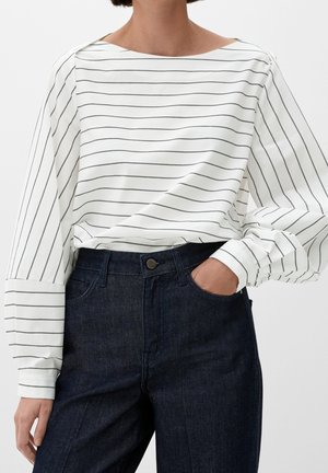 Personne portant un chemisier blanc à manches longues avec de fines rayures horizontales noires rentré dans un jean bleu foncé taille haute, une main dans la poche.