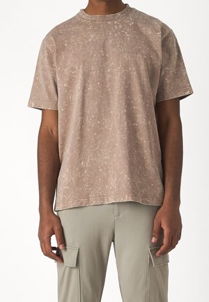 Uomo che indossa una t-shirt marrone sbiadita a maniche corte con un piccolo logo "BOSS" sul petto e pantaloni cargo grigio chiaro.