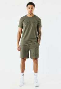 T-shirt en coton vert olive avec du texte en script noir, associé à un short assorti comportant le même texte. Des chaussettes blanches et des baskets complètent la tenue.