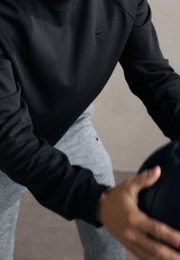 Personne portant une chemise noire à manches longues et un pantalon gris tenant un ballon d'exercice noir, penchée en avant dans un cadre intérieur.