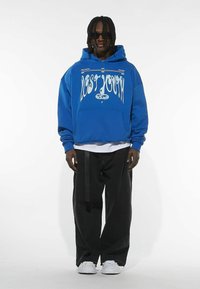 Blå oversized hoodie med hvid "LOST YOUTH" grafik, kenguru lomme, lagdelt med en hvid skjorte, parret med sorte vide bukser og hvide sneakers.