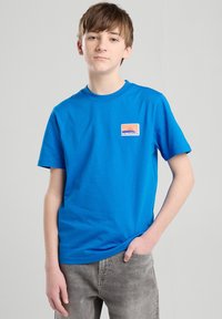 Sininen puuvillapaita, jossa on pyöreä kaula-aukko ja lyhyet hihat, sekä oranssi-valkoinen California-logo. Yhdistetty harmaisiin housuihin.