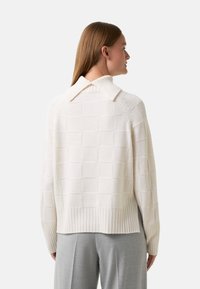Vrouw met steil bruin haar draagt een crèmekleurige gebreide trui met een brede ribbelrand en een omgeslagen kraag, gecombineerd met lichtgrijze pantalon.