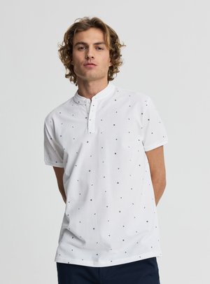 Giovane uomo con capelli ricci che indossa una camicia bianca a maniche corte con piccoli motivi a croce neri, in piedi contro uno sfondo chiaro e uniforme.