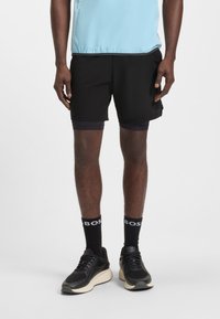 Muž v černých běžeckých šortkách s vrstveným designem, černých ponožkách BOSS a černých teniskách s béžovými podrážkami, stojící před bílým pozadím.