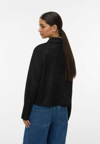 Veste noire à texture lisse, col classique et coupe courte. Portée sur un jean bleu, avec une coupe décontractée et des poches arrière.