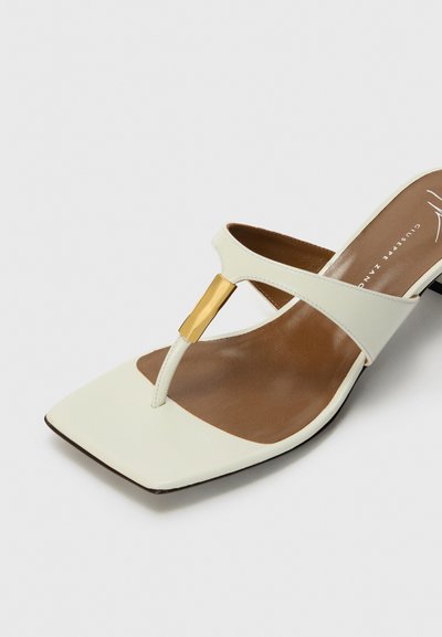Sandelă din piele albă cu vârf pătrat, având un accent vertical aurit și o branț de culoare maronie. Bretele subțiri creează un design minimalist.