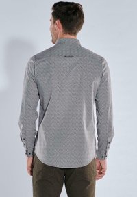 Mann mit kurzen braunen Haaren, der ein langärmliges, gemustertes Hemd in Hellgrau und olivgrüne Hosen trägt, von hinten vor einem schlichten Hintergrund.