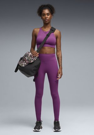 Frau in lila Sport-BH und Leggings hält eine schwarze Sporttasche mit Blumenmotiv, trägt schwarze Sneakers und steht vor grauem Hintergrund.