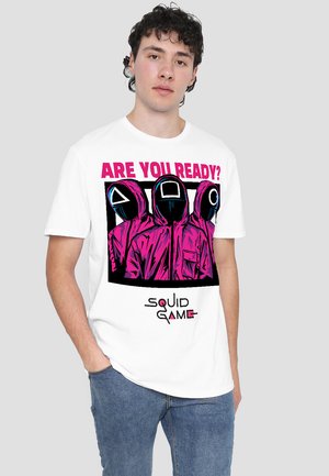 Weißes Baumwoll-T-Shirt mit einem auffälligen Grafikdruck maskierter Figuren in pinken Umhängen, mit dem Text "BIST DU BEREIT?" und "SQUID GAME" darunter.