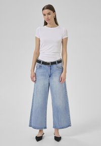 Witte korte mouwen shirt gecombineerd met lichtblauwe wijde jeans en een zwarte riem. Kenmerken zijn een rechte zoom en minimale slijtage. Zwarte instapschoenen.