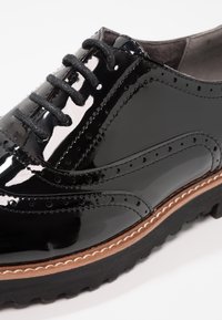 Chaussure en cuir verni noir avec un design à lacets, présentant des détails en brogue, une texture lisse, et une semelle contrastante en cuir tan avec des rainures.