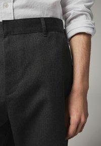 Pantaloni din denim negru cu o textură netedă și o croială ajustată. Talie plată cu un detaliu discret de cusătură. Mâna se odihnește pe lateral.