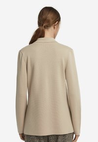 Beige blazer i texturerad stickad kvalitet, med slagkrage och långa ärmar. Slät yta med rak skärning bak.