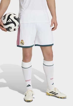 Osoba ubrana w białe krótkie spodenki piłkarskie Real Madrid, białe skarpetki z logo RMCF oraz korki, trzyma czarno-białą piłkę do piłki nożnej Adidas.