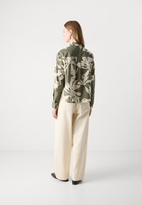 Chemise verte à manches longues avec des motifs floraux blancs, à col, associée à un pantalon crème ample et des chaussures noires. Texture lisse, coupe décontractée.