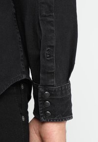 Manica di camicia in denim nero con chiusura a bottone, caratterizzata da un design elegante, cuciture visibili e tre bottoni a pressione sul polsino.