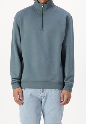 Lätthanterlig, zippered pullover i dämpad teal, med hög krage och ribbade ärmslut. Mjuk textur, med avslappnad passform och minimal detaljer.