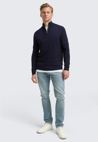 Mann, der einen marineblauen Viertelreißverschluss-Pullover, ein weißes Unterhemd, hellblaue Jeans und weiße Sneaker trägt und vor einem einfarbigen Hintergrund steht.
