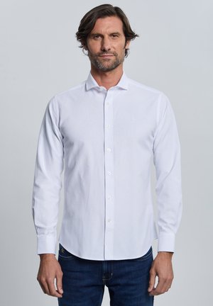 Hombre con cabello oscuro y barba que lleva una camisa blanca de manga larga abotonada y vaqueros azul oscuro, de pie frente a un fondo claro y liso.
