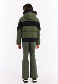 Olijfgroene pufferjack met zwarte horizontale strepen, voorzien van een capuchon, gequilte structuur, en gecombineerd met bijpassende groene uitlopende broek en zwarte laarzen.
