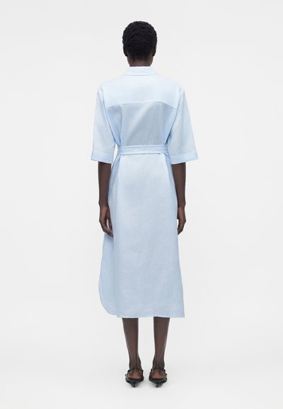 Γυναίκα που στέκεται πλάτη φοράει ένα ανοιχτό μπλε μίντι φόρεμα με ζώνη, μανίκια μέχρι τους αγκώνες και μαύρα σανδάλια με τακούνι.