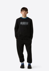 Svart hettegenser med grå tekstlogo, kombinert med svarte joggebukser. Har rund hals og ribbestrikkede mansjetter. Stilfullt satt sammen med blå og svarte sko og mønstrede sokker.