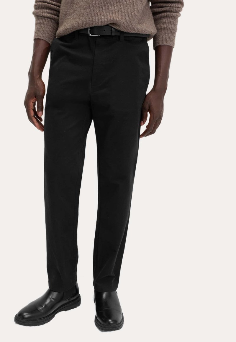 Homme portant un pantalon noir sur mesure, une ceinture noire, des chaussures en cuir noires et un pull marron tricoté, debout devant un fond clair.