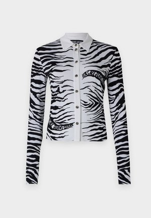 Majica z dezenom zebre v črno-beli barvi z ovratnikom, zapiranjem na gumb in dolgimi rokavi. Na sprednji strani je napis z logotipom.