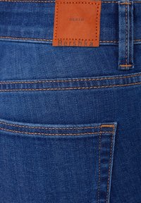 Vaqueros de mezclilla azules con costuras naranjas y una etiqueta de cuero marrón etiquetada "DENIM Bershka". Presenta un bolsillo trasero y una textura suave.