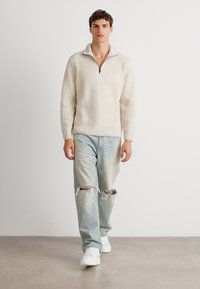 Helles Beige, gerippter Strickpullover mit einem Viertel-Reißverschluss-Kragen, kombiniert mit leicht blauen, distressed Jeans und weißen Sneakers. Einfaches Design.