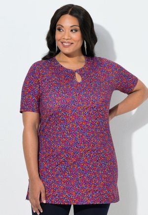 Femme souriante aux longs cheveux foncés portant des boucles d'oreilles pendantes et une tunique courte à manches courtes violette à motif floral avec une encolure en goutte d'eau.