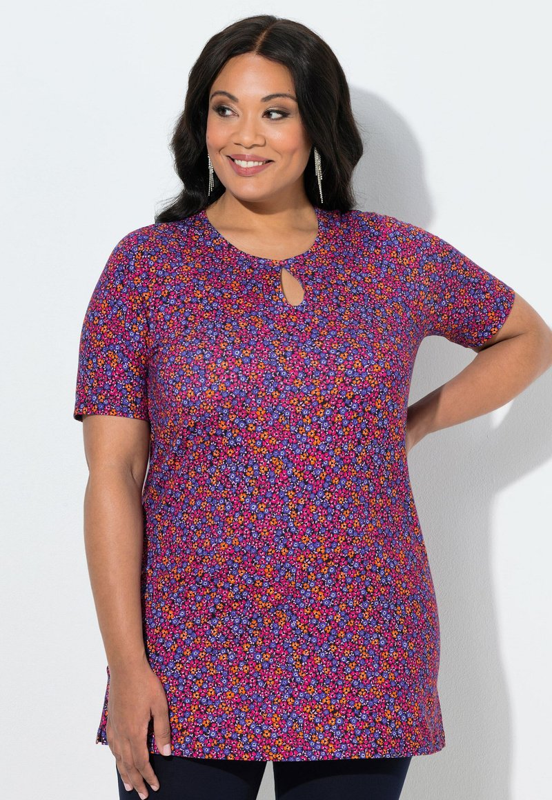 Mujer sonriente con cabello largo y oscuro, que lleva pendientes colgantes y una túnica morada de flores con mangas cortas y un escote en forma de ojo de cerradura.