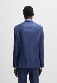 Giacca elegante blu con texture, con uno spacco singolo sul retro, polsini con bottoni e linee pulite, abbinata a pantaloni coordinati.