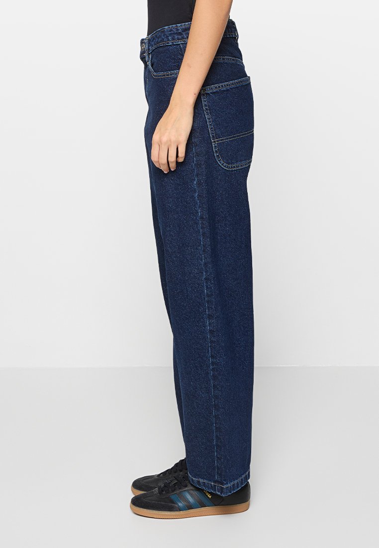Jeans a gamba larga in denim blu scuro con cuciture visibili, una tasca posteriore e vita alta, abbinati a sneaker nere con strisce blu.
