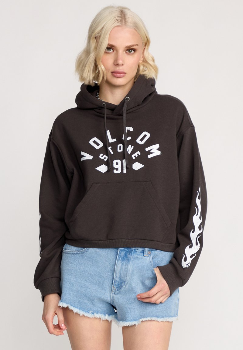 Haut à capuche noir court avec le texte blanc "VOLCOM" et des graphismes de flammes sur les manches, associé à un short en denim déchiré bleu clair.