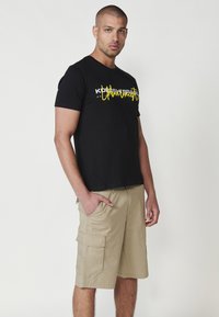 Camiseta de algodón negra con texto gráfico amarillo y acentos blancos, combinada con pantalones cortos de cargo beige que tienen bolsillos laterales y un corte relajado.
