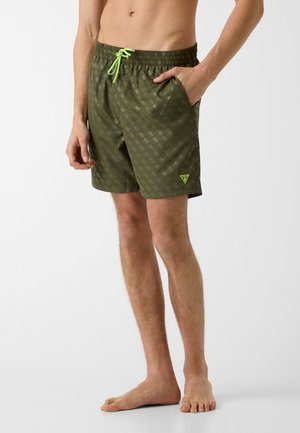 Männliches Model trägt olivgrüne Badeshorts mit neonfarbenem Kordelzug und dezentem Muster, steht barfuß mit einer Hand in der Tasche.