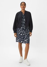 Veste aviateur navy avec des manches côtelées sur une robe midi à motif abstrait blanc, associée à des baskets blanches.